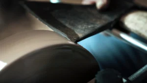 metal tool being sharpened on a grinding wheel. The tool, possibly a chisel or plane blade, is held against the rotating wheel, creating sparks and shaping the edge.